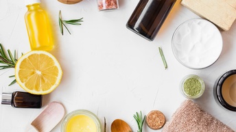 Verschiedene Objekte zum Thema Kosmetik wie Honig, braune Fläschchen, grünes Pulver, ein kleiner Holzlöffel, ein Handtuch, gelbe Flüssigkeit in einem durchsichtigem Fläschchen sind in einem Kreis auf weißem Hintergrund angeordnet.