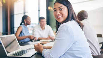 Eine Person in einem hellem Oberteil sitzt an einem Tisch, blickt direkt in die Kamera und hat vor sich einen aufgeklappten Laptop. Im Hintergrund sitzen drei Personen am selben Tisch und reden miteinander