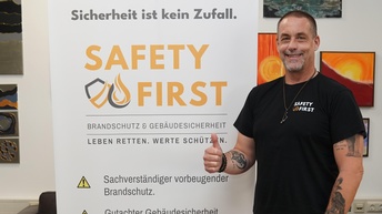 Ein Mann steht lächelnd neben einem großen Roll-up-Banner mit der Aufschrift „SAFETY FIRST“. Er trägt ein schwarzes T-Shirt mit demselben Logo und zeigt den Daumen nach oben. Auf dem Banner steht: „Sicherheit ist kein Zufall. SAFETY FIRST – Brandschutz & Gebäudesicherheit. Leben retten. Werte schützen.“ Darunter folgen Informationen zu Sachverständigenleistungen im vorbeugenden Brandschutz, Gutachten nach ÖNORMEN B1300/B1301 sowie Schulungen. Im Hintergrund hängen mehrere bunte Gemälde an der Wand