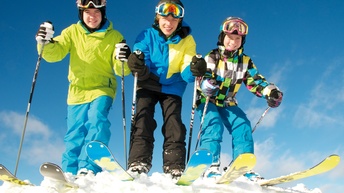 Drei jugendliche Personen in Schianzügen mit Schi, Schistöcken und Helmen mit Schibrillen lächeln auf Schnee stehend unter blauem Himmel