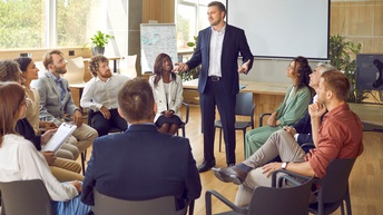 In einem hellen Raum sitzen mehrere Personen in Bürokleidung in einem Sesselkreis. Eine der Personen steht und spricht zu den anderen. Im Hintergrund sind ein beschriebenes Whiteboard und eine Leinwand