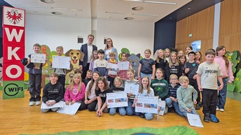 Eine große Gruppe von rund 25 Kindern steht gemeinsam mit zwei erwachsenen Begleitpersonen in einem Veranstaltungsraum der Wirtschaftskammer. Die Kinder halten Arbeitsblätter in der Hand und schauen in die Kamera. Im Hintergrund sind bunte Tierfiguren und ein „WKO“-Aufsteller zu sehen.