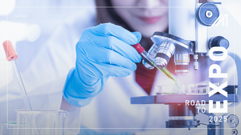 Nahaufnahme von einer Person, die blaue Schutzhandschuhe trägt und mit einer Pipette eine grüne Flüssigkeit in ein Schälchen unter ein Mikroskop gibt