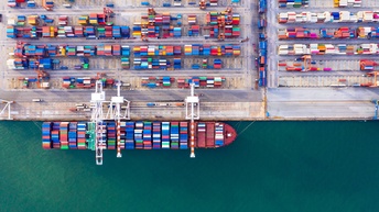 Top-Shot eines großen Schiffs mit vielen bunten Frachtcontainern an Deck, das auf einer Seite von einem Gewässer umgeben ist und auf der anderen Seite von einer asphaltierten Fläche, auf der weitere bunte Container sind.