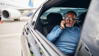 Person mit Brillen sitzt auf Rückbank eines Autos und telefoniert, im Hintergrund verschwommen Flugzeug