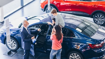 Drei Personen stehen neben einem Auto. Das Auto steht in einem Raum mit einer Fensterfront. Eine Person hält die Fahrertür auf. Auf den Schultern einer anderen Person sitzt ein Kind. Neben dem Auto steht ein weiteres Auto