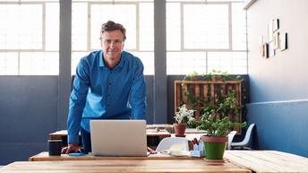 Fescher Unternehmer steht lächelnd vor seinem Schreibtisch mit Laptop