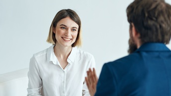 Person mit kurzen Haaren und weißer Bluse sitzt und hält ein Klemmbrett sowie einen Stift und blickt auf eine Person in Unschärfe ihr gegenüber