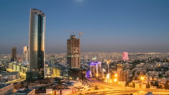 Panoramaaufnahme von Amman bei Nacht mit Wolkenkratzern sowie mehrspurigen Autostraßen im Vordergrund. Dahinter erstrecken sich viele Gebäude