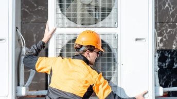 Rückenansicht einer Person mit schwarz-oranger Jacke, orangem Helm und Sonnenbrille, die neben einem Außengerät einer Klimaanlage steht und beide Hände auf dem Gerät hat
