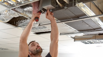 Person mit Bart hält Bohrmaschine Kopfüber an Lüftungsgerät