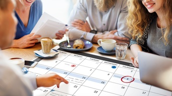 Mehrere lächelnde Personen sitzen um Tisch und blicken auf einen ausgebreiteten Terminkalender.