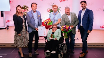 6 Menschen vor WKW-Banner, Gratulation, Blumen, Mensch im Rollstuhl