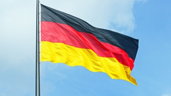 An einer Metallstange ist die Flagge Deutschlands montiert. Die Flagge weht im Wind. Dahinter ist der blaue Himmel mit einigen weißen Wolken