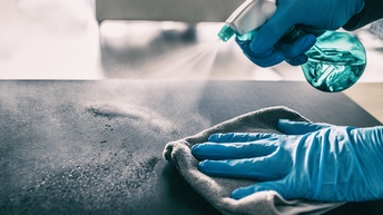 Nahaufnahme der Hände einer Person mit Handschuhen, die einen Lappen hält und mit einer Sprühflasche Flüssigkeit auf einem Möbelstück mit glatter Oberfläche verteilt