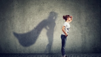 Person mit langen dunklen geschlossenen Haaren mit Brille steht mit aufrechter Haltung und blickt in die Ferne während an der Wand ein Schatten der Person mit einem Superhero-Cape erscheint