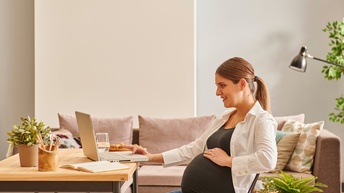 Schwangere Person sitzt freudig bei einem Wohnzimmertisch und arbeitet mit einem Laptop während im Hintergrund eine Couch sowie Pflanzen stehen