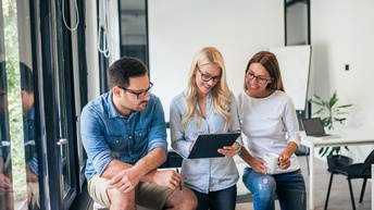 Drei Personen stehen nebeneinander. Die mittlere Person hält einen Notizblock, auf den alle blicken. Im Hintergrund ist ein Whiteboard