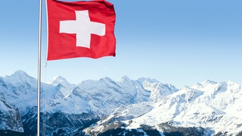 In der linken Bildhälfte ist die Schweizer Flagge an einer Metallstange montiert. Die Flagge weht im Wind. Im Hintergrund sind schneebedeckte Berge. Der Himmel ist blau