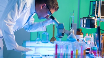 Eine Person in einem Arbeitskittel ist seitlich fotografiert. Sie blickt durch ein Mikroskop, das auf einem Tisch aus Glas steht. Auf diesem befinden sich auch Pipetten und Reagenzgläser mit Flüssigkeiten, sowie Lampen.