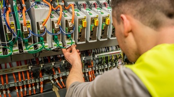 Rückenansicht einer Person, die sich nach vorne beugt und ihre linke Hand auf einem grauen, sehr kleinen Kästchen hat, auf dem ein grüner Zettel klebt. Um dieses Kästchen sind viele weitere sowie viele unterschiedliche Kabel.