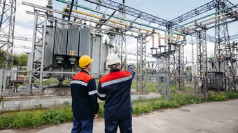 Zwei Personen in Arbeitskleidung und Schutzhelmen stehen vor einem umzäunten Umspannwerk. Eine Person hebt den Arm und zeigt auf Bauteile der Anlage. Im Hintergrund sind Transformatoren, Leitungen, Isolatoren und Metallkonstruktionen zu sehen