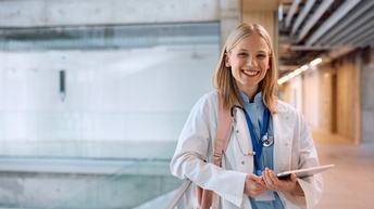 Eine lächelnde Person steht in einem Gebäude und hält ein Tablet in den Händen. Sie trägt einen weißen Kittel und hat ein Stethoskop um den Hals.