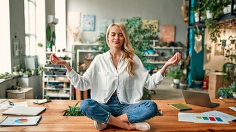 Eine Person in Jeans, Turnschuhen, weißer Bluse sowie mit offenen, hellen Haaren sitzt in einem Büroraum mit grünen Pflanzen auf einem großen Tisch und bildet mit den Händen jeweils ein Mudra