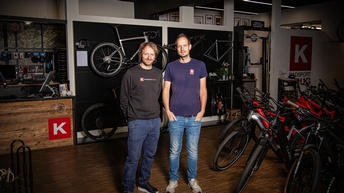 Die Aufnahme zeigt zwei Unternehmer in ihrem Fahrradfachgeschäft. Sie stehen im Verkaufsraum zwischen mehreren Fahrrädern und E-Bikes. Hinter ihnen befinden sich an der Wand präsentierte Rennräder, Fahrradrahmen und Zubehör. Rechts ist ein Bereich mit E-Bikes zu sehen, im Hintergrund eine Werkstatt für Service- und Reparaturarbeiten. Die moderne Einrichtung vermittelt den Eindruck eines spezialisierten Fachbetriebs für Verkauf, Wartung und individuelle Beratung im Radsportbereich.