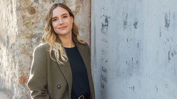 Porträt einer lächelnden Person mit hellen, offenen Haaren, dunklem Oberteil sowie Sakko darüber, die neben einer Mauer steht