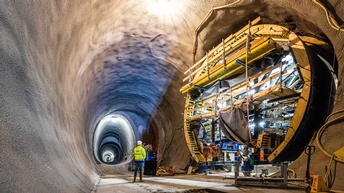In einem großen Tunnel steht eine Person mit Schutzweste und Helm. Rechts neben der Person ist ein sehr großes Gerät zur Bearbeitung von Tunneln