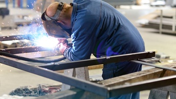 Eine Person in Rückenansicht in blauem Overall mit Schutzvisier schweißt ein Metallgerüst.