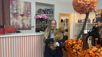 Porträt einer Person in einem Sakko und langen, offenen, hellen Haaren, die in einem Geschäft steht und einen Hund hält