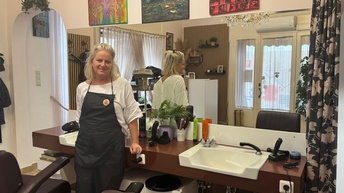 Nahaufnahme einer Person mit blonden Haaren und einer Schürze, die in einem Friseursalon steht. Hinter ihr ist eine Anrichte mit einem eingefassten Waschbecken und Friseurprodukten wie Shampoos sowie ein großer Spiegel, der die Rückenansicht der Person und den Eingangsbereich des Friseursalons spiegelt