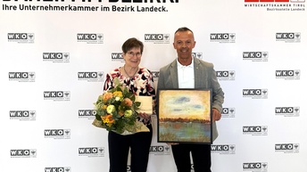 Zwei Personen stehen vor einer WKO-Tirol-Fotowand im Bezirk Landeck. Eine Frau hält einen Blumenstrauß und ein Kuvert, ein Mann präsentiert ein gerahmtes Landschaftsbild. Beide schauen in die Kamera.