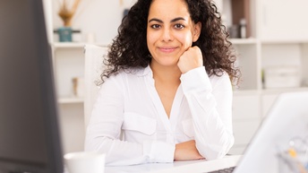 Portrait einer Person mit dunklem lockigen Haar und weißer Bluse, die sich mit ihrer Hand bei einem Schreibtisch abstützt, daneben steht ein Monitor sowie ein Laptop und ein Kaffeebecher auf dem Tisch und dahinter zeigt sich ein weißes Regal