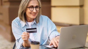 Lächelnde Person mit Brillen Bistrotisch vor aufgeklappten Laptop sitzend hält Bankkarte in Händen und blickt auf Monitor, auf Tisch verschwommen im Vordergrund Kaffeebecher