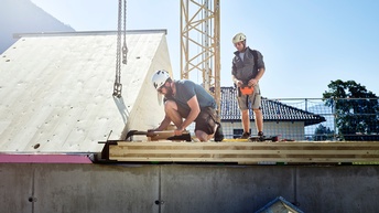 Zwei Personen bei der Montage von Holzbrettern am Dach, im Hintergrund blauer Himmel und gelber Kran