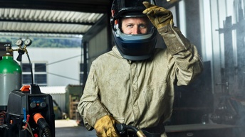 Ein Mann in Schutzausrüstung mit Schweißgerät hebt das Visier seines Helms und lächelt direkt in die Kamera