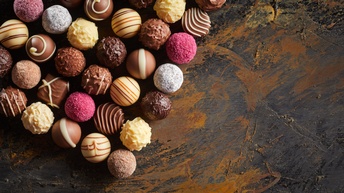 Topshot vieler, kleiner, runder Pralinen, die auf einer dunklen Oberfläche liegen. Manche Pralinen sind dunkel, andere hell, andere rosa. Manche haben Striche oben, manche sind uneben