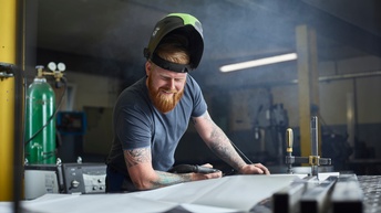 Ein Mann mit hochgezogenem Schweißerhelm kontrolliert einen Plan, der vor ihm auf dem Tisch ausgebreitet liegt