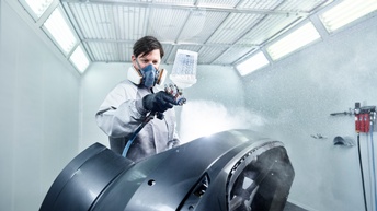 Ein Karosseriebautechniker sprüht ein Autoteil mit einer Flüssigkeit ein. Er trägt eine Atemschutzmaske.