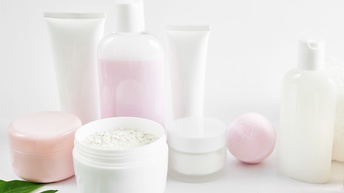 Verschiedene Verpackungen für Kosmetika wie Tuben, Tiegel oder Fläschchen aus Plastik ohne Etikett, ein Tiegel mit Puder ist geöffnet, eine rosa Badekugel sowie Blätter liegen daneben