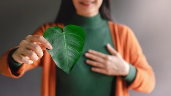 Fokus auf grünes Blatt, das Person in Hand hält, die im Hintergrund verschwommen im Ausschnitt erkennbar ist und eine Hand auf ihr Herz hält