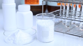 Auf einem hellen Tisch stehen nebeneinander zwei Glasgefäße. In beiden ist ein weißes Pulver drinnen. Auf dem rechten steht L-Lysine. Dahinter stehen weiße, kleine Plastikgefäße