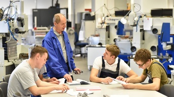 Stehende Person in blauer Arbeitskluft spricht zu sitzenden jungen Personen, die Unterlagen sichten, im Hintergrund technische Gerätschaften