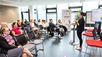 Person in Seitenansicht steht auf einer Bühne und spricht in ein Mikrofon einem sitzenden Publikum zugewandt, im Hintergrund Rollup mit Schriftzug Vielfalt vernetzt