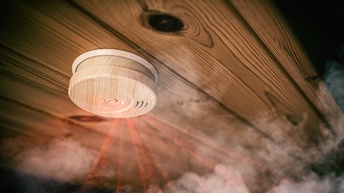Nahaufnahme eines Rauchmelders, der an einer Holzdecke montiert ist. Vom Rauchmelder gehen rote Lichter weg. Um den Rauchmelder ist Rauch