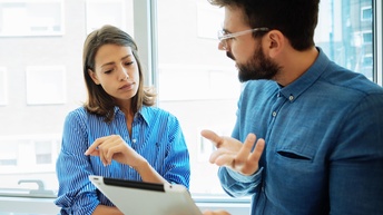 Eine Person mit kurzen Haaren, Bart und Brille hält ein Tablet in Richtung einer weiteren Person mit der sie zugewandt spricht, die zweite Person blickt interessiert auf das Tablet, im Hintergrund zeigt sich eine vollflächige Glasfront