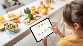 Person hat Obst und Gemüse vor sich am Tisch liegen und hält ein Tablet in der Hand. Am Tablet sieht man einen Onlineshop für Speisen.
